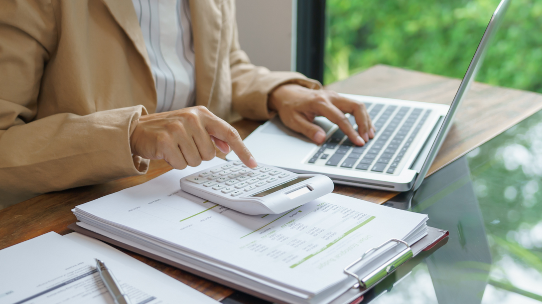 You can see a woman sitting at a desk. You can not see her head. you can only see her from her shoulders down. You would be looking at her from the right of her. she is wearing a tan long sleeve jacket with a white and blue striped shirt underneath. she has her left hand on top of an open tablet keyboard her thumb is on the spacebar and  her four other fingers  are resting on the keys above the spacebar. her right hand into a fist with index finger pointing at the calculator. There is a stack of papers with a binder clipped to them underneath the calculator. the laptop is to the left side of the calculator and then on the right side there is another stack of papers with a silver pen laying on top of it. the desk underneath the paperwork and laptop is half dark wood and half glass. Behind her is a wall half of it is a window. Outside of the window is blurred green shrubs but bright like the sun is out.